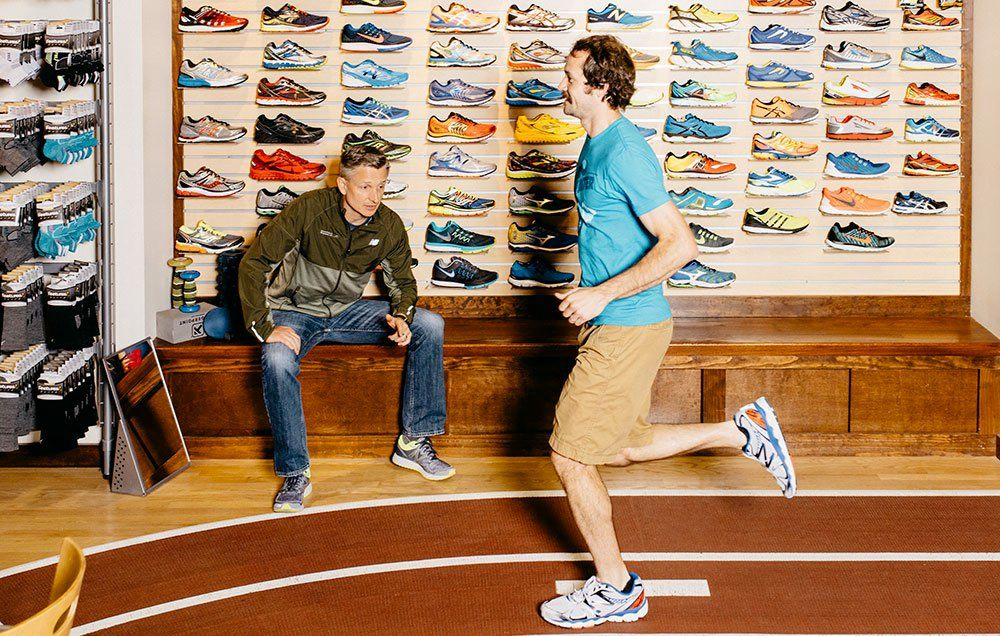A man in a shoe store trying shoes and running while another man sitting on the bench looking 